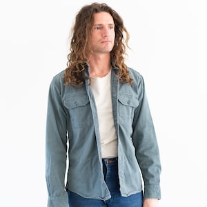 May include: A man with long, curly brown hair is wearing a blue long-sleeved shirt over a white t-shirt and blue jeans.