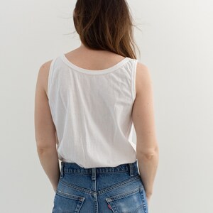 May include: A woman wearing a white tank top and blue denim jeans. The jeans have a faded look and are high-waisted.