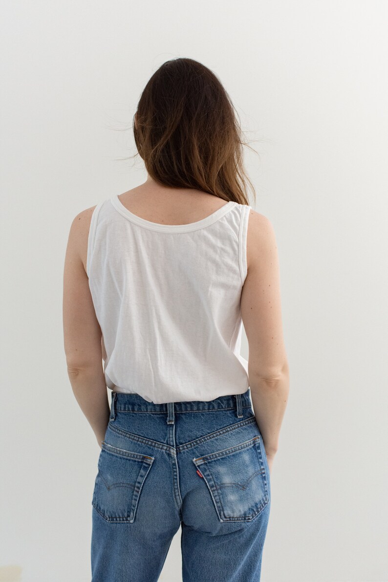 May include: A woman wearing a white tank top and blue denim jeans. The jeans have a faded look and are high-waisted.