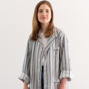 May include: A gray and white striped jacket with a pocket on the chest and rolled-up sleeves. The jacket is buttoned and worn over a white t-shirt and blue jeans. The jacket has a classic, relaxed fit.