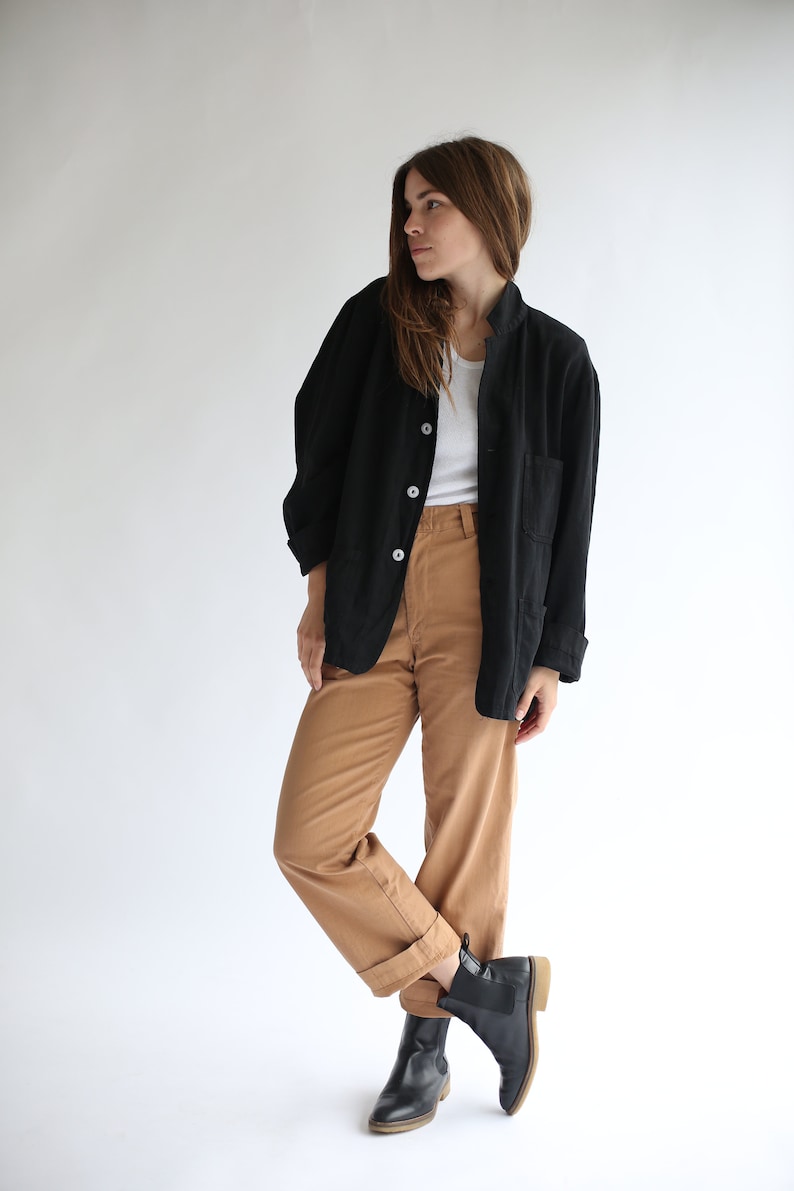 May include: A woman wearing a black jacket over a white shirt and brown trousers. She is wearing black ankle boots.