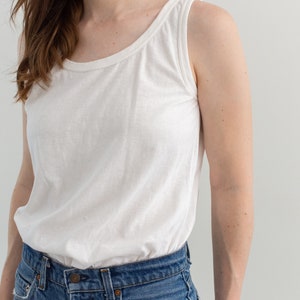 May include: A white tank top with a scoop neck and a relaxed fit. The tank top is paired with blue denim jeans.