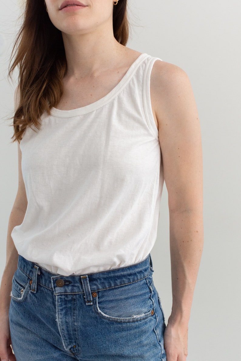 May include: A white tank top with a scoop neck and a relaxed fit. The tank top is paired with blue denim jeans.