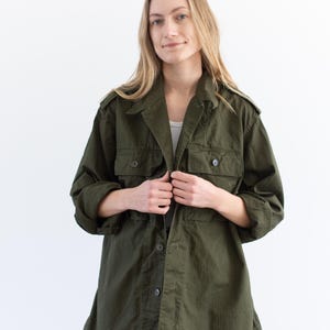 May include: An olive green utility jacket featuring a collared neckline, long sleeves, and two button-flap chest pockets. The jacket has a button-down front closure with dark buttons and shoulder epaulets, worn over a white shirt and blue jeans.
