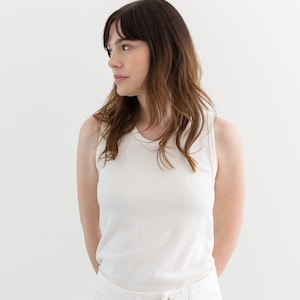 May include: A woman wearing a white sleeveless shirt and white pants. She is standing in front of a white background.