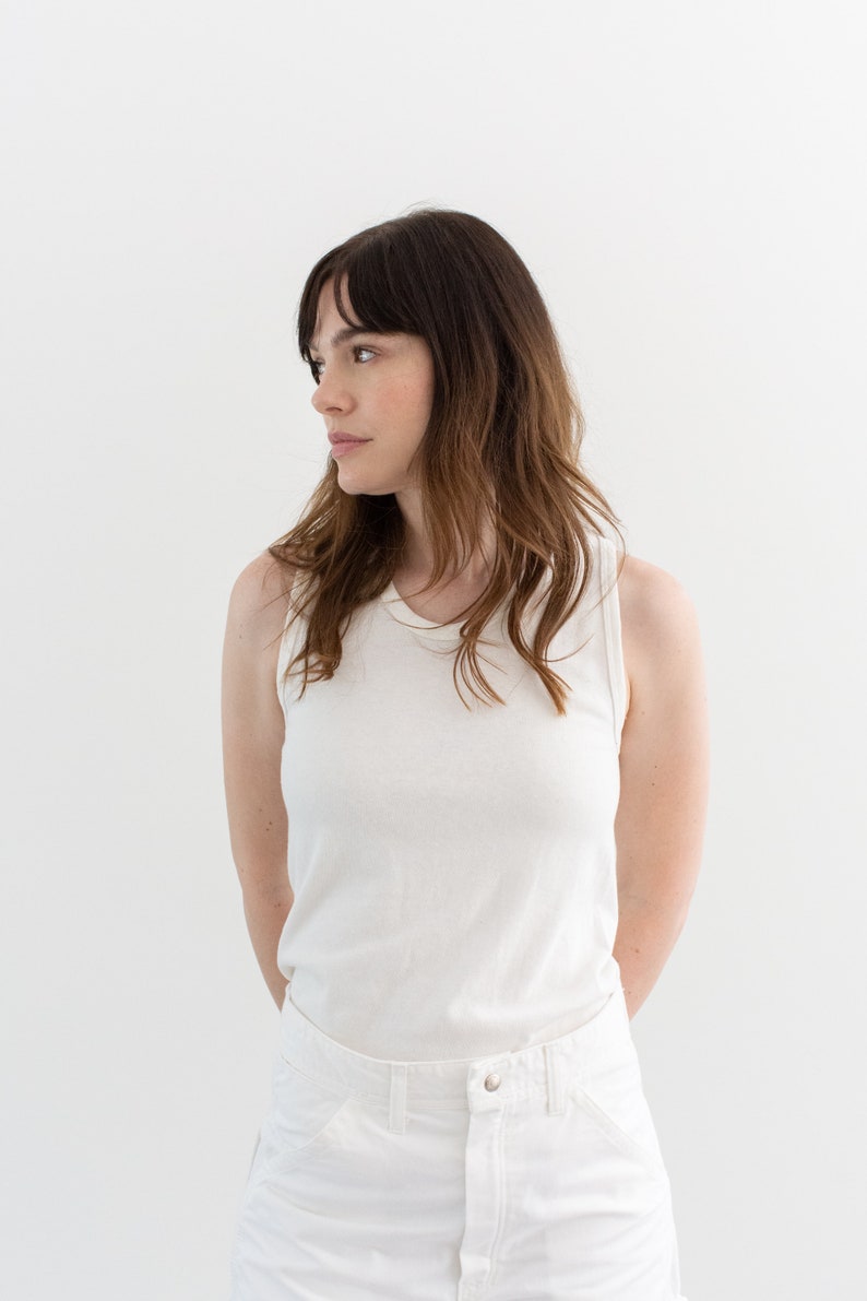 May include: A woman wearing a white sleeveless shirt and white pants. She is standing in front of a white background.
