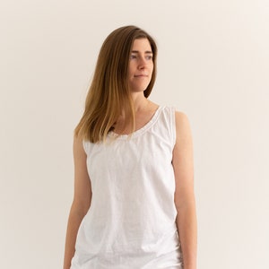 May include: A woman wearing a white tank top and blue jeans. She is standing in front of a white wall.