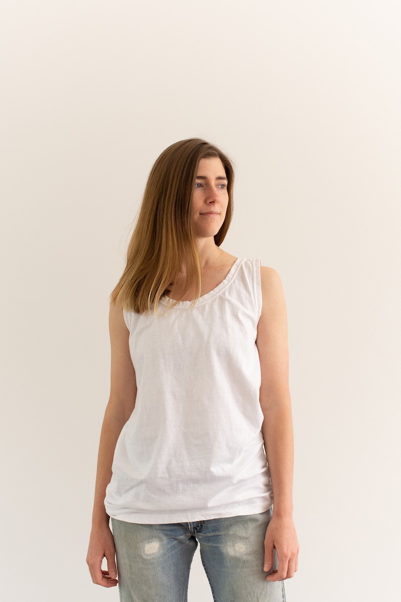May include: A woman wearing a white tank top and blue jeans. She is standing in front of a white wall.