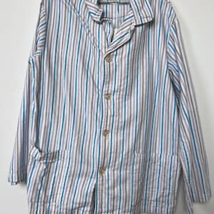 Op de afbeelding: Een gestreept overhemd in wit, blauw en grijs, met een kraag en knoopsluiting. Het shirt met lange mouwen heeft twee zakken en hangt aan een zwarte hanger. De verticale strepen creëren een klassiek patroon.