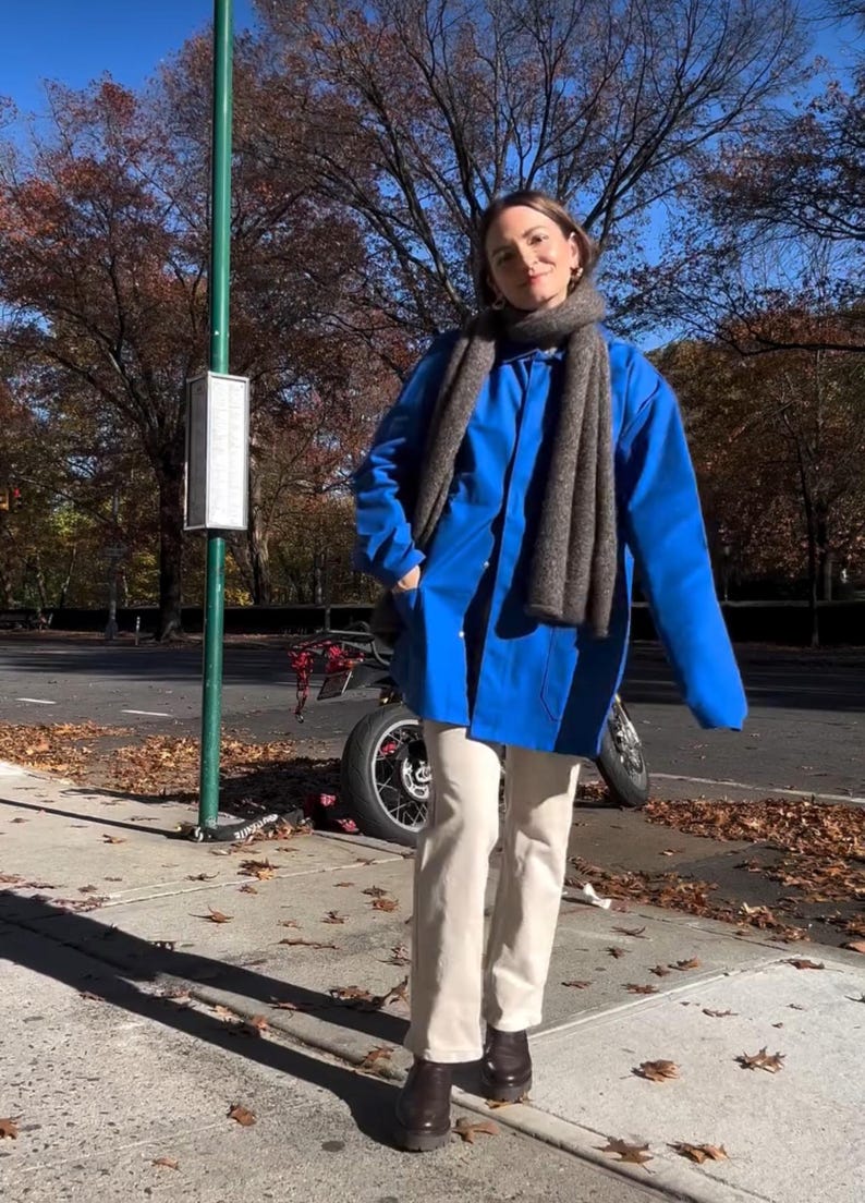 May include: A woman wearing a blue jacket with a gray scarf, beige pants, and brown boots. She is standing on a sidewalk with fallen leaves.