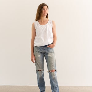 May include: A woman wearing a white tank top and light blue denim jeans with distressed details. She is standing on a concrete floor in front of a white wall.