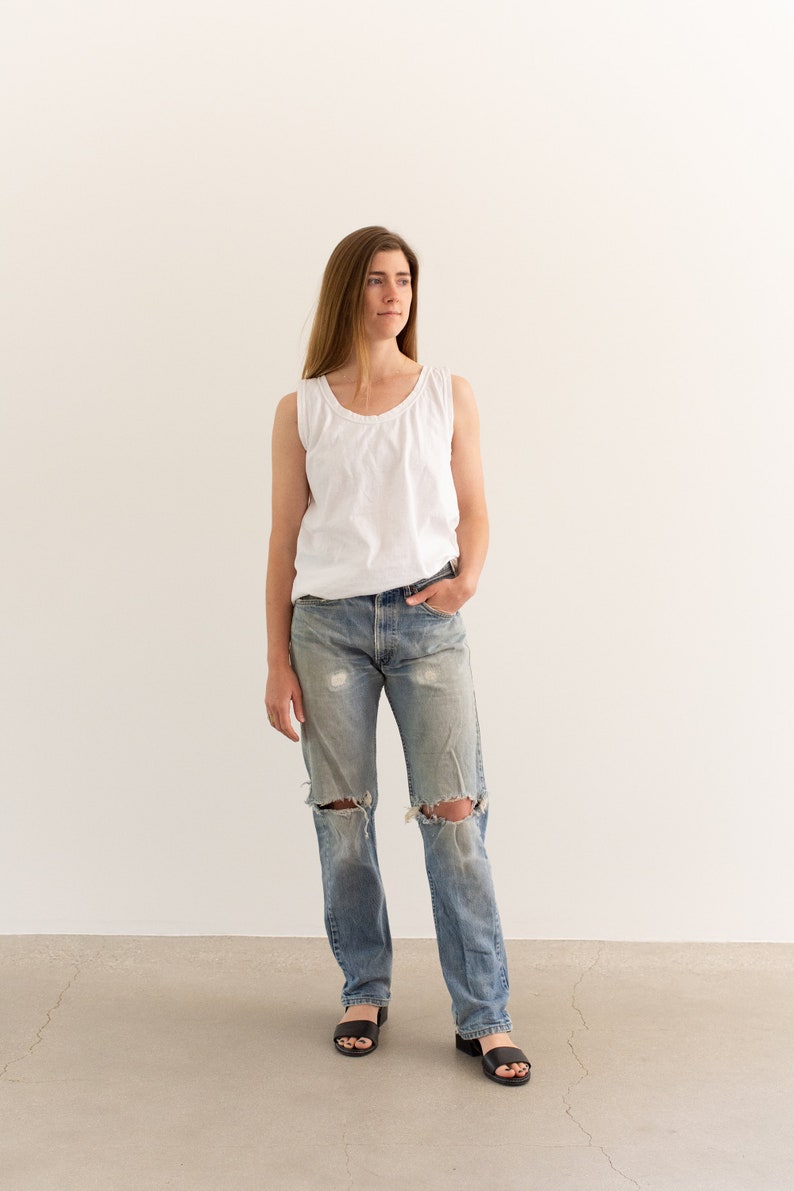 May include: A woman wearing a white tank top and light blue denim jeans with distressed details. She is standing on a concrete floor in front of a white wall.