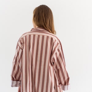 May include: A long-sleeved, oversized shirt with vertical stripes in shades of brown, white, and burgundy. The cuffs are rolled up, revealing a contrasting pattern. The shirt is worn with blue jeans.