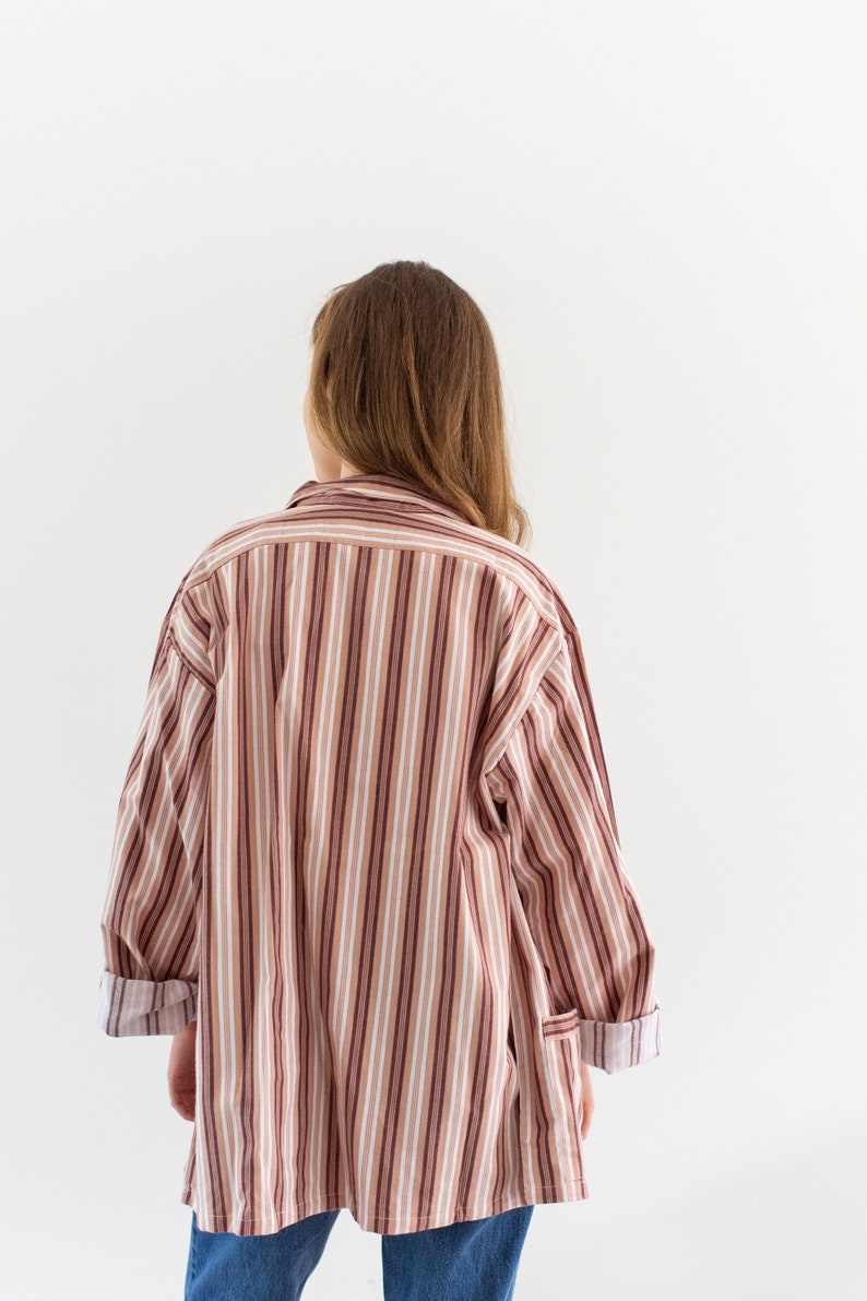 May include: A long-sleeved, oversized shirt with vertical stripes in shades of brown, white, and burgundy. The cuffs are rolled up, revealing a contrasting pattern. The shirt is worn with blue jeans.