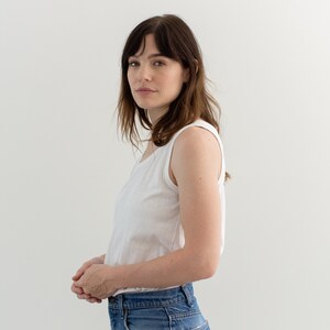May include: A woman wearing a white sleeveless top and blue jeans. She is standing in front of a white wall.