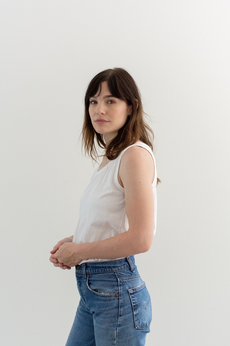May include: A woman wearing a white sleeveless top and blue jeans. She is standing in front of a white wall.