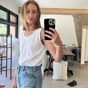 May include: A woman wearing a white sleeveless shirt and light blue denim jeans. She is taking a selfie with a black smartphone.