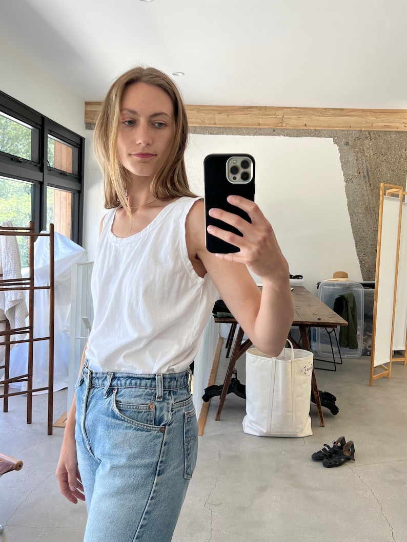 May include: A woman wearing a white sleeveless shirt and light blue denim jeans. She is taking a selfie with a black smartphone.