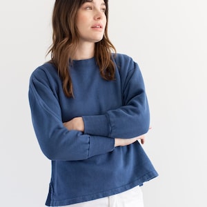 May include: A woman wearing a blue long-sleeved sweatshirt with a relaxed fit and white pants.
