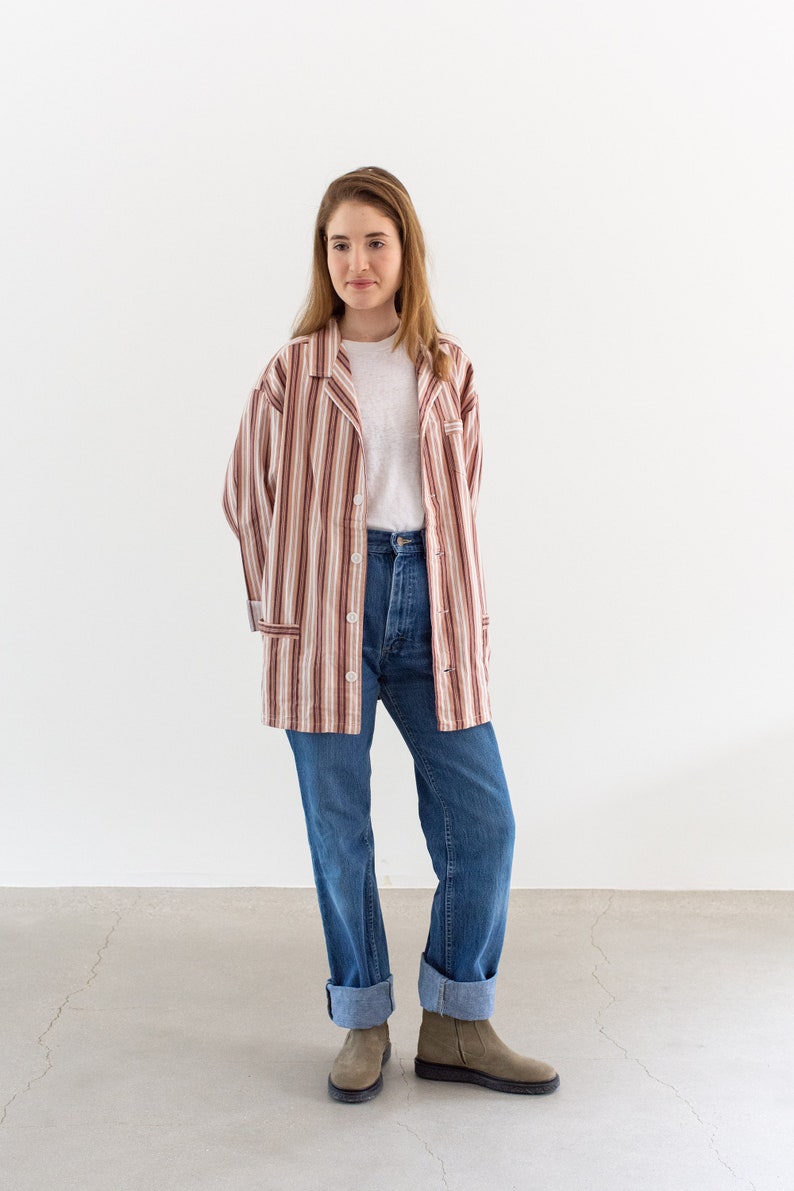 May include: A long-sleeved, button-down shirt with vertical stripes in shades of brown, tan, and white. The shirt is paired with a white t-shirt, blue jeans, and gray ankle boots. The jeans have rolled-up cuffs.
