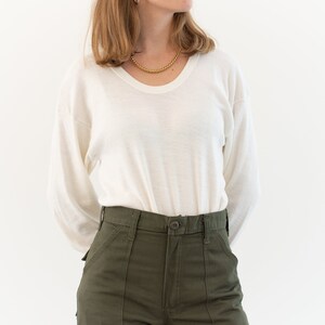 May include: A long-sleeve white crew neck shirt with a thin, lightweight texture paired with olive green utility pants featuring a high waist, a button closure, and two large front patch pockets. The shirt is tucked into the pants.
