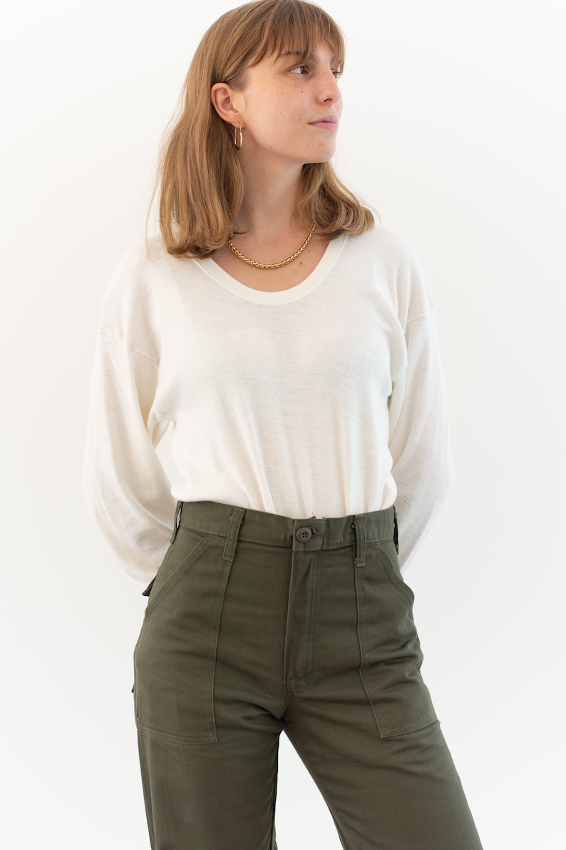 May include: A long-sleeve white crew neck shirt with a thin, lightweight texture paired with olive green utility pants featuring a high waist, a button closure, and two large front patch pockets. The shirt is tucked into the pants.