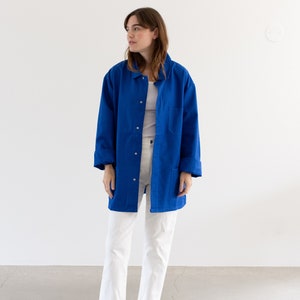 May include: A woman wearing a long, royal blue work jacket over a white t-shirt and white pants. She is wearing black sandals.