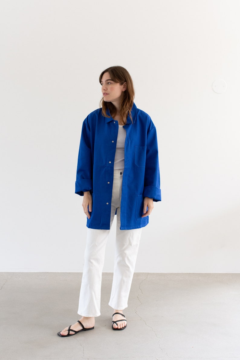 May include: A woman wearing a long, royal blue work jacket over a white t-shirt and white pants. She is wearing black sandals.