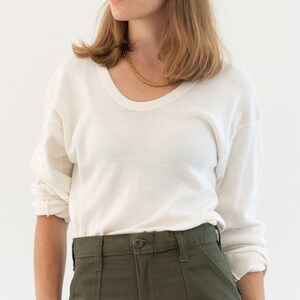 May include: A long-sleeve off-white sweater with a scoop neckline and dropped shoulders, paired with olive green trousers featuring a front button closure and visible belt loops. A gold-toned chain necklace rests around the neck.