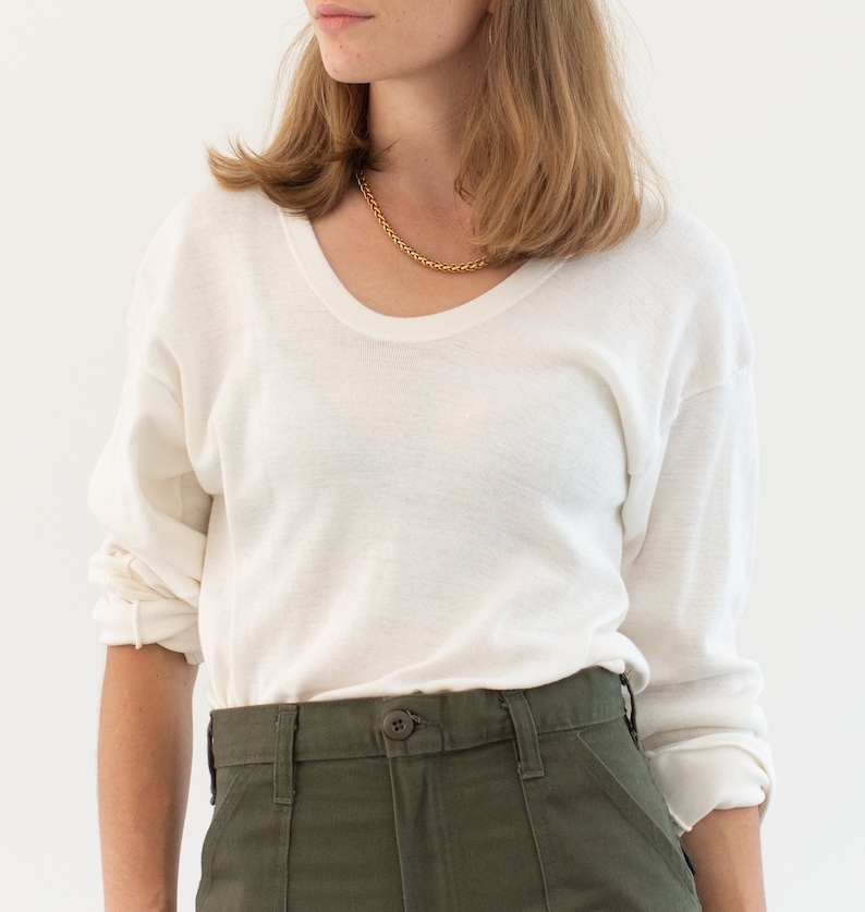 May include: A long-sleeve off-white sweater with a scoop neckline and dropped shoulders, paired with olive green trousers featuring a front button closure and visible belt loops. A gold-toned chain necklace rests around the neck.
