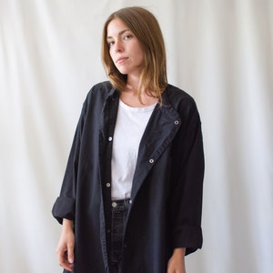 May include: A black long-sleeved shirt with a button-up front and a collar. The shirt is worn over a white t-shirt and black jeans.