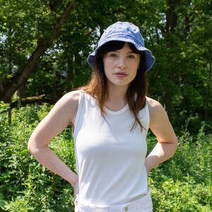 May include: A woman wearing a white tank top and white shorts with a blue denim bucket hat. She is standing in a grassy area with trees in the background.
