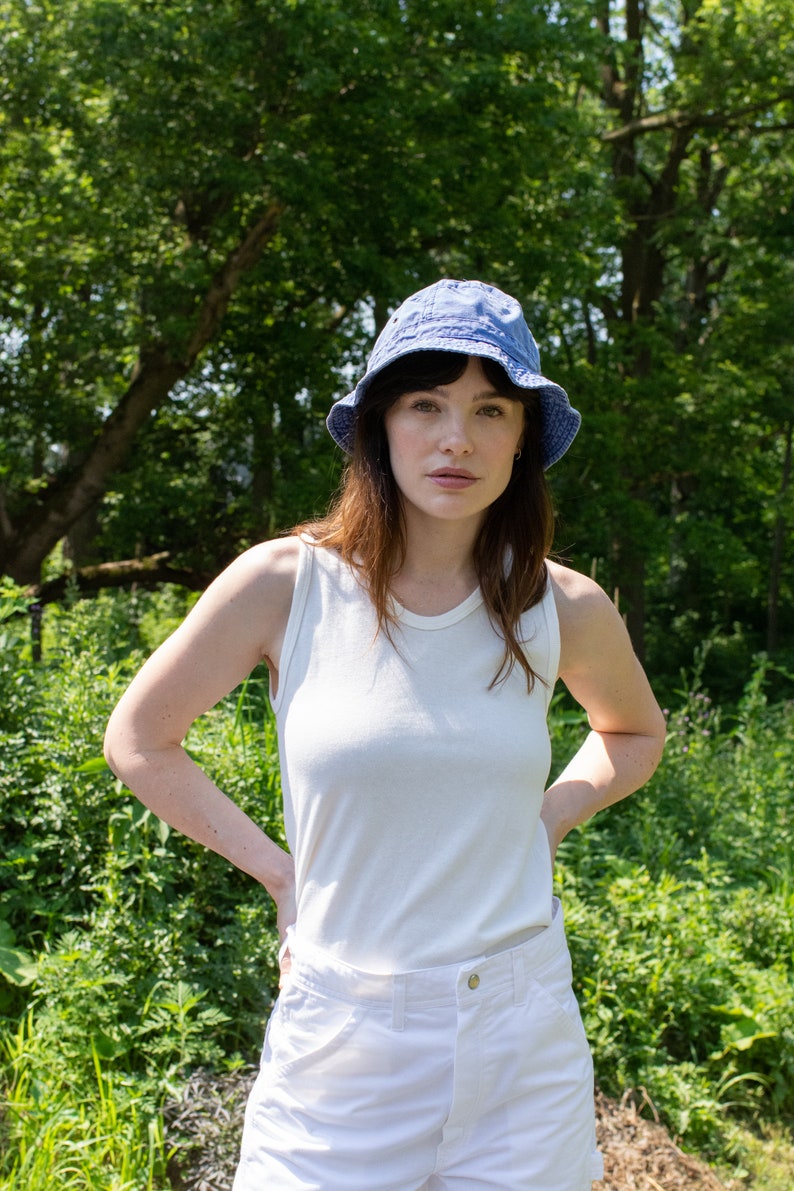 May include: A woman wearing a white tank top and white shorts with a blue denim bucket hat. She is standing in a grassy area with trees in the background.