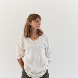 May include: A long-sleeved, off-white crew neck sweater with a relaxed fit, paired with olive green trousers featuring front patch pockets. The sweater has a lightweight, slightly sheer texture and is worn with a gold-toned chain necklace.