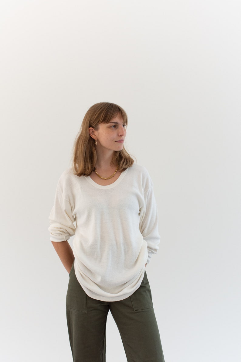 May include: A long-sleeved, off-white crew neck sweater with a relaxed fit, paired with olive green trousers featuring front patch pockets. The sweater has a lightweight, slightly sheer texture and is worn with a gold-toned chain necklace.