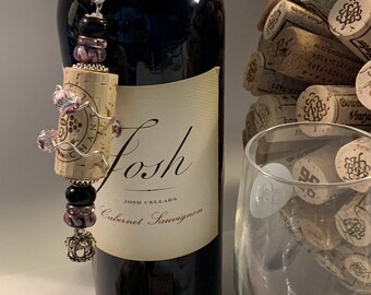 Op de afbeelding: Een donkere wijnfles met een decoratieve kurk en een kralenbedel. De fles is voorzien van het label "Josh Cabernet Sauvignon". Een wijnglas en een stapel kurken zijn ook zichtbaar.