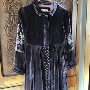 May include: A dark grey velvet jacket with embroidered floral details on the sleeves and a beaded trim down the front. The jacket has a collared neckline and a button closure.