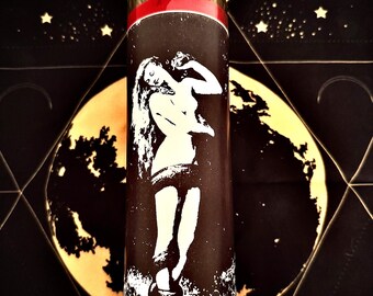 May include: A tall, cylindrical candle with a black background featuring a white illustration of a woman. The candle has a red base and a gold-colored top with a silver crescent moon and star design. The background is black with gold celestial patterns.