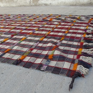 May include: A handwoven rug with a checkered pattern in shades of red, white, gray, and orange. The rug has fringe along the edges.