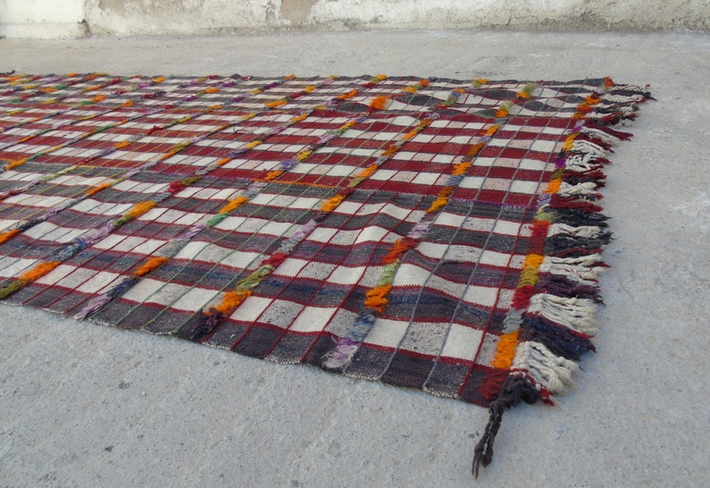 May include: A handwoven rug with a checkered pattern in shades of red, white, gray, and orange. The rug has fringe along the edges.