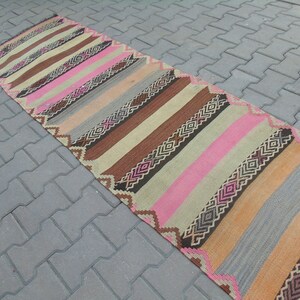 May include: A long, narrow rug with a striped pattern in shades of brown, pink, beige, and gray. The rug has a geometric design with diamond shapes.