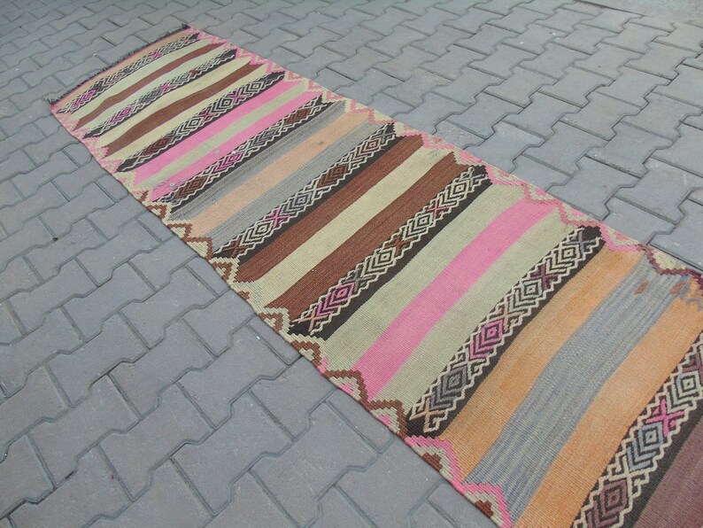 May include: A long, narrow rug with a striped pattern in shades of brown, pink, beige, and gray. The rug has a geometric design with diamond shapes.