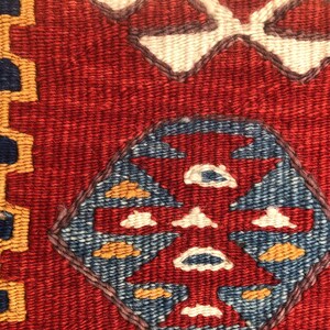 May include: Close-up of a woven textile with a red background and geometric patterns. The design includes blue, white, and yellow shapes, with a border of blue and gold. The texture is visible, suggesting a handmade rug or fabric.