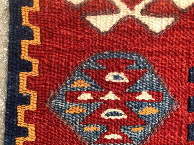 May include: Close-up of a woven textile with a red background and geometric patterns. The design includes blue, white, and yellow shapes, with a border of blue and gold. The texture is visible, suggesting a handmade rug or fabric.