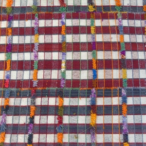 May include: A close-up of a woven fabric with a checkered pattern in red, white, and gray. The fabric has a fringe border with colorful threads.
