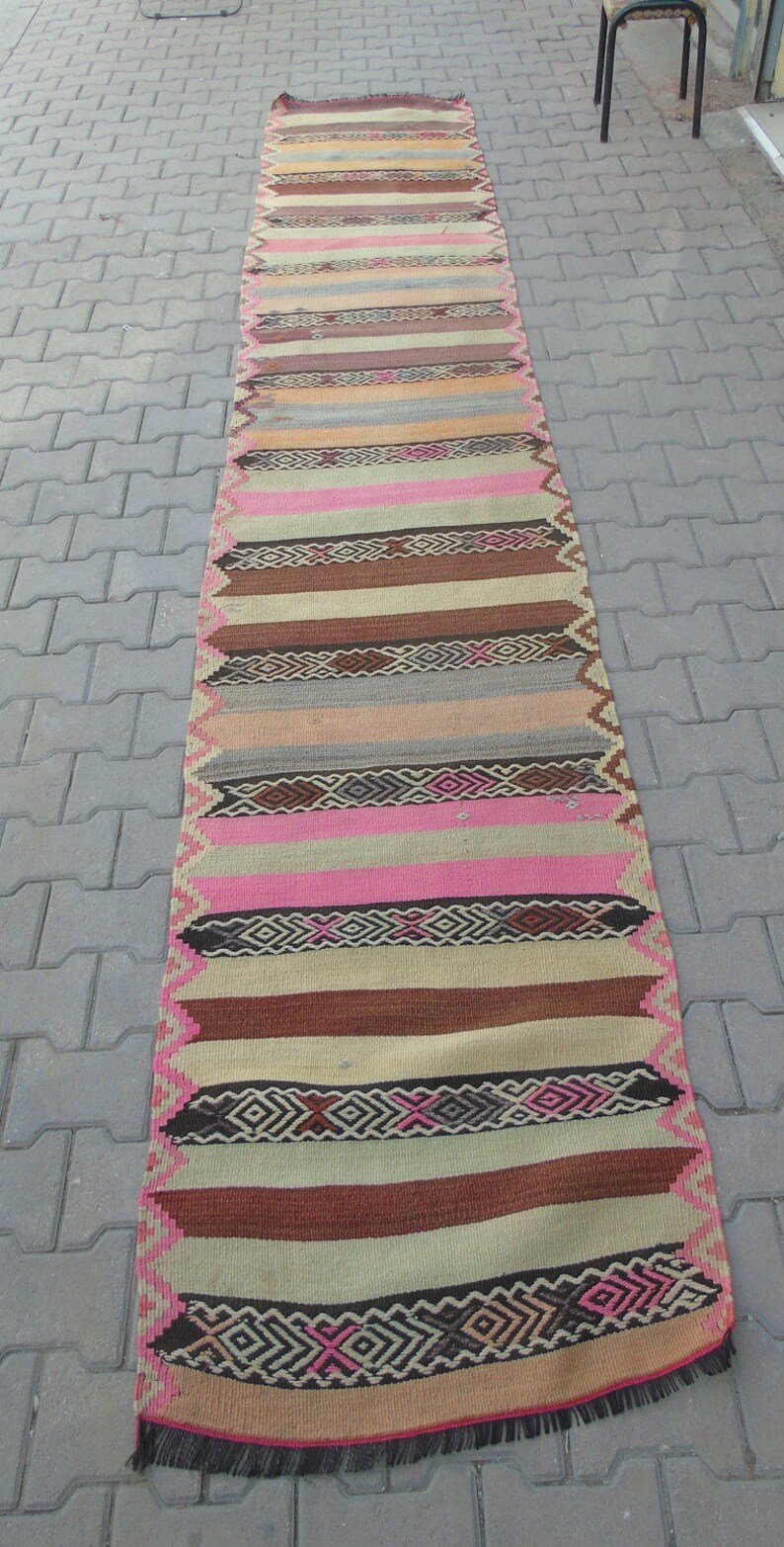 May include: A long, narrow rug with a colorful striped pattern. The rug has a fringe on both ends and is made of woven fabric. The colors in the rug include pink, brown, beige, and black.