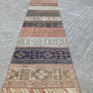 Può includere: Un tappeto lungo e stretto con un design a patchwork. Il tappeto è composto da diverse strisce di tessuto, ognuna con un motivo unico. I colori del tappeto sono per lo più tenui, con sfumature di marrone, beige e rosa.