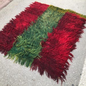 May include: A rectangular rug with a shaggy texture, featuring three vertical stripes in shades of red and green. The rug is made of a thick, plush material, and the edges are slightly frayed. The rug is suitable for a living room.