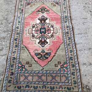 May include: A vintage pink and green rug with a geometric pattern and floral accents. The rug has a fringe border and is approximately 2 feet by 4 feet.