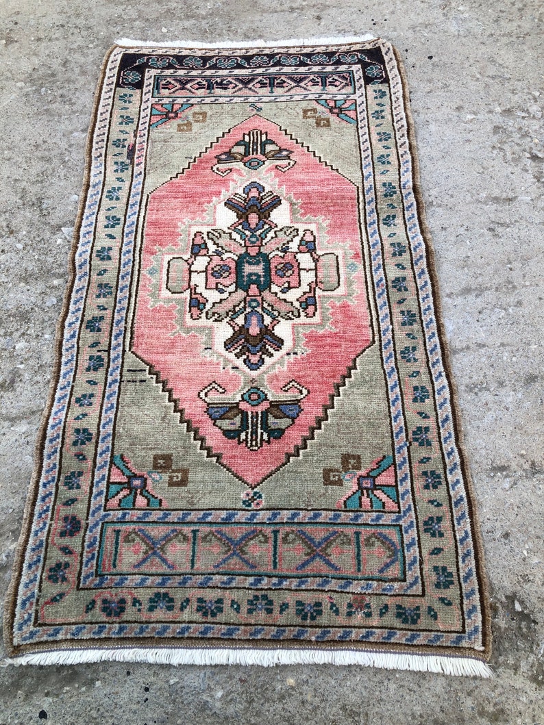 May include: A vintage pink and green rug with a geometric pattern and floral accents. The rug has a fringe border and is approximately 2 feet by 4 feet.